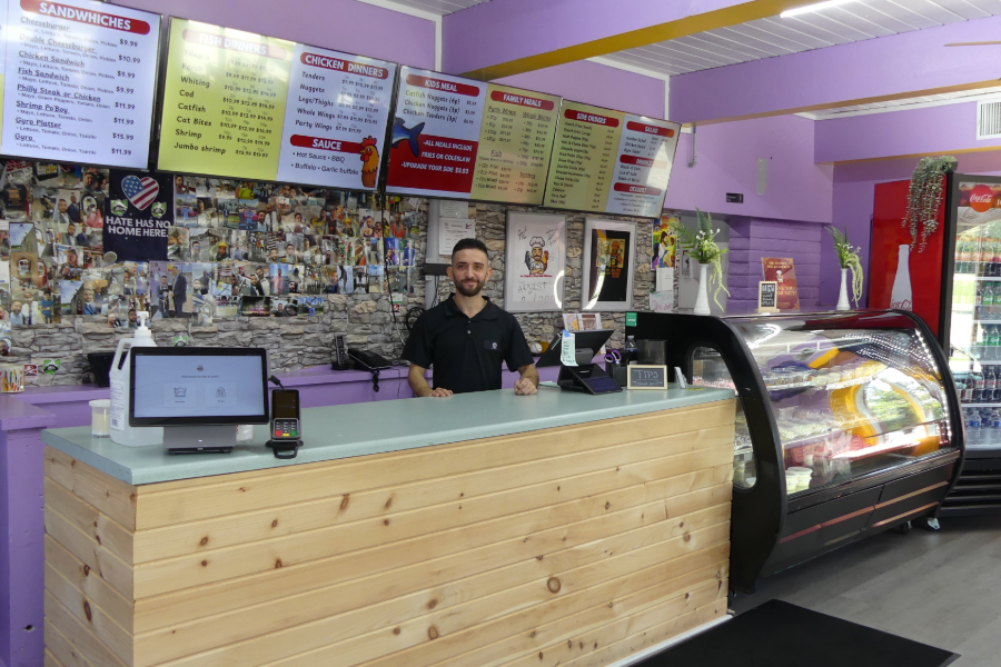 Moe Alhayek, owner of Mr. Hayek's Fish & Chicken, smiling behind the counter with the menu boards and a Hate Has No Home Here sign on the wall